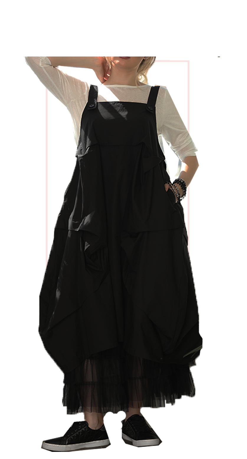 A woman wearing a black oversized bib-style dress over a white long-sleeve top, paired with black sneakers, shown against a plain white background.