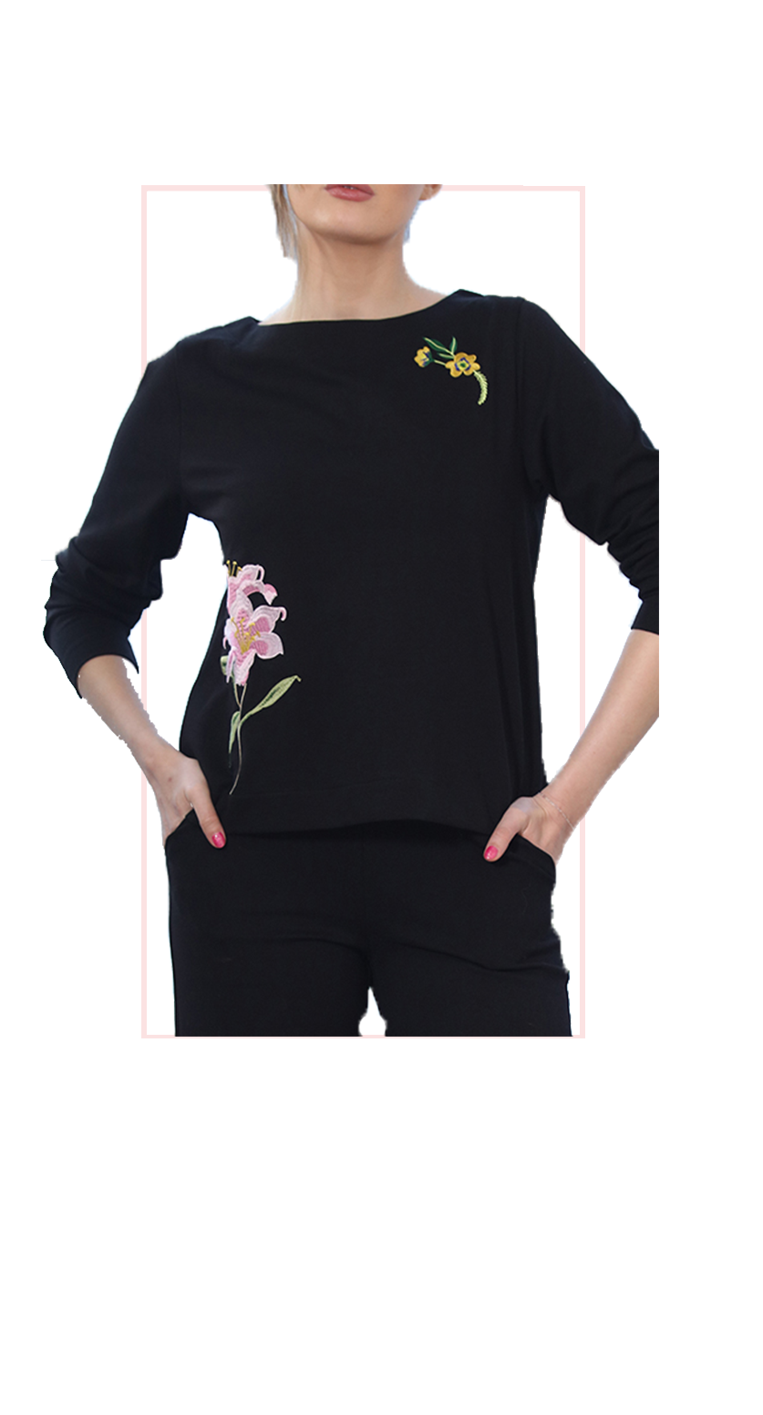 A woman wearing a black long-sleeve top with pink and yellow floral embroidery on the front, paired with black pants, shown against a plain white background.